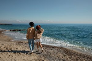Couple in hats walking along a sandy beach with blue sea and clear sky background