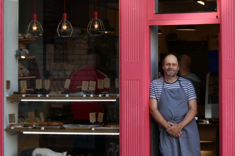 Bakery owner standing in red-framed doorway, wearing apron, with pastries displayed inside under modern pendant lights