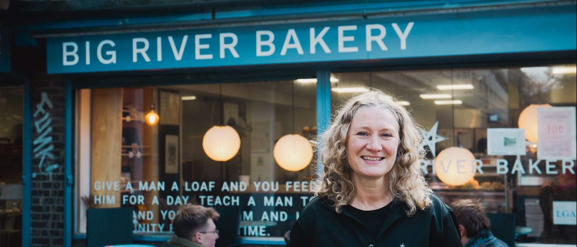 Person smiling in front of Big River Bakery shopfront, offering fresh bread and community spirit