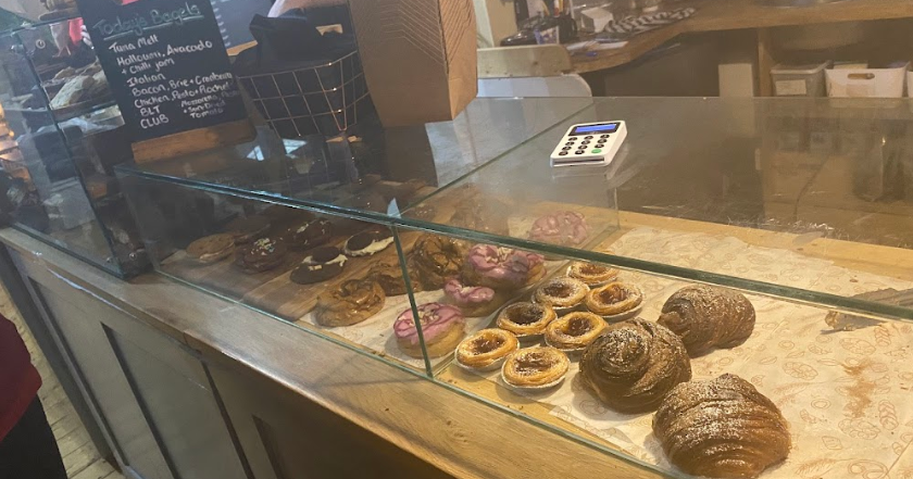 Bakery counter display with assorted pastries and bagels behind glass, including croissants and tarts, in a cosy shop setting