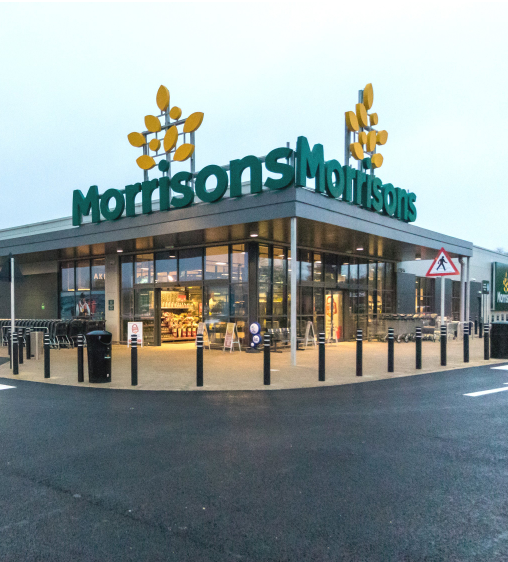 Morrisons supermarket front entrance with signs and trolleys in the foreground, large logo visible on the building facade