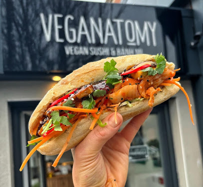 Hand holding vegan banh mi sandwich with fresh vegetables, showcased in front of Veganatomy shop sign