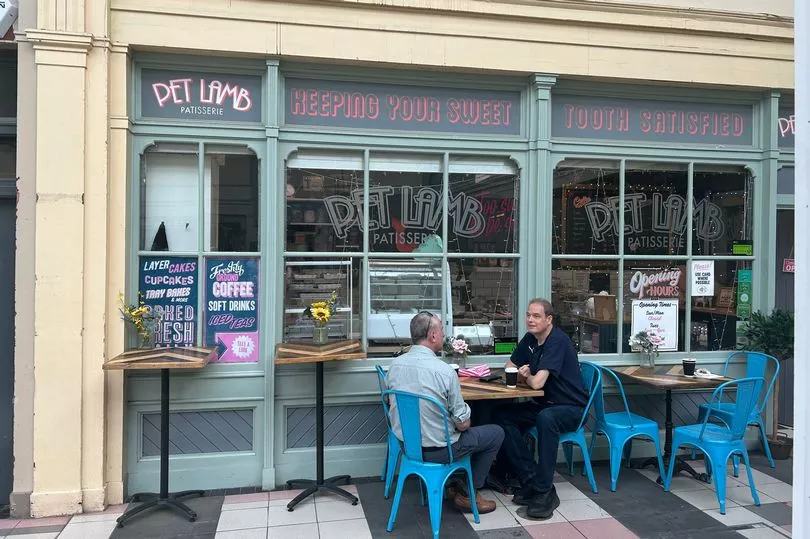 Two men seated outdoors at a patisserie café with blue chairs and tables decorated with flowers