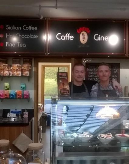 Two smiling café staff at Caffè Ginevra counter with Italian coffee and desserts on display