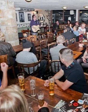 People enjoy live guitar performance in a cosy pub setting with drinks on wooden tables